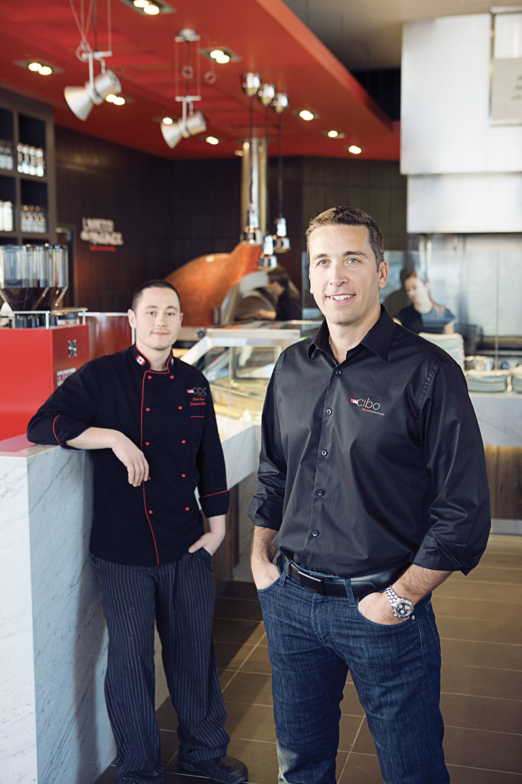 Two male restaurant staff smiling in a modern kitchen, one in a black chef coat leaning on a marble counter and the other in a black shirt with hands in his pockets.