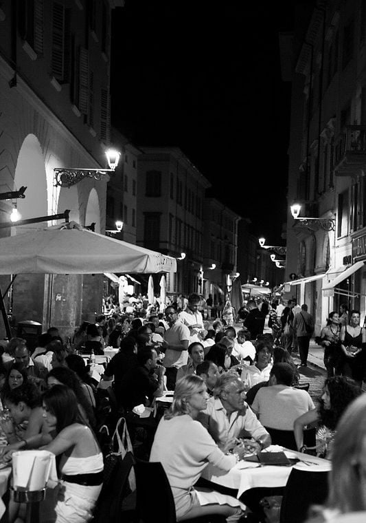 Crowded outdoor restaurant terrace on a narrow European street at night with diners, umbrellas, and street lamps in black and white