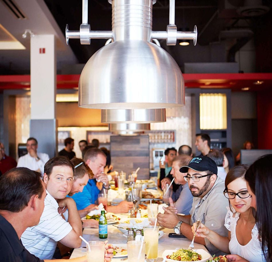 Busy restaurant scene with diners sharing a long table under large metal pendant lights, eating and chatting.