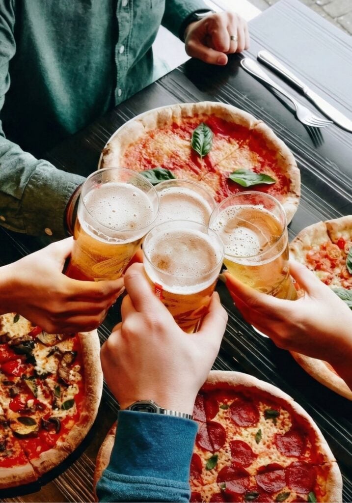 Four hands clinking beer glasses over a table filled with several pizzas (pepperoni, margherita and veggie).