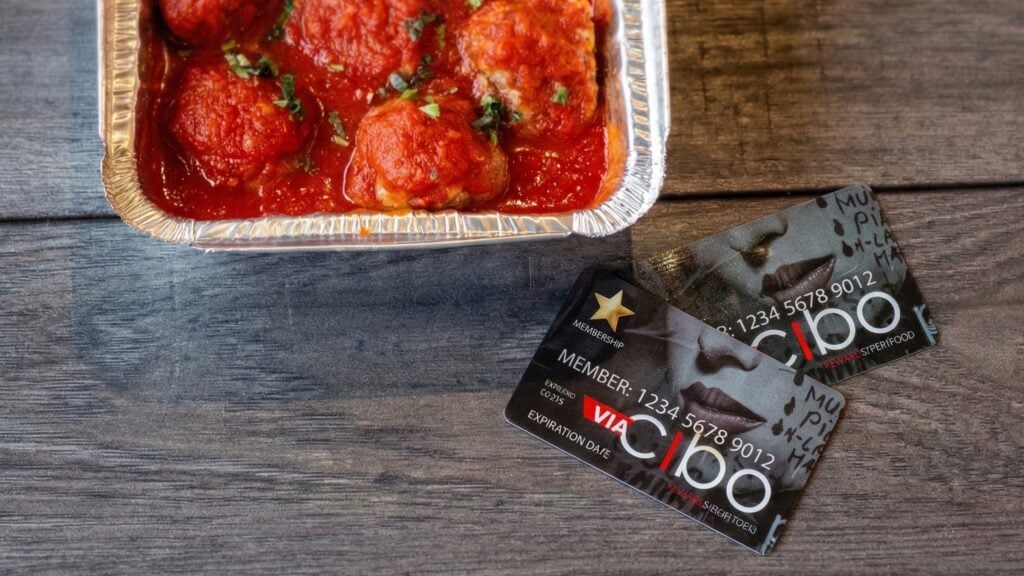 Aluminum tray of tomato-covered meatballs on a wooden table next to two black membership/rewards cards partially overlapping