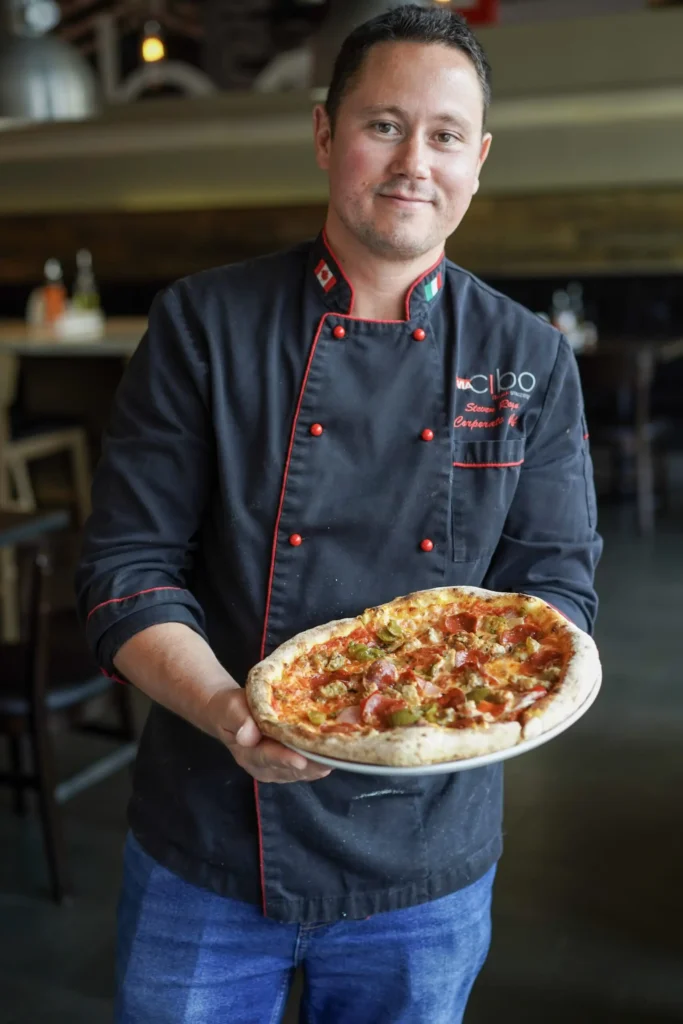 Smiling chef in a black jacket with red trim holding a freshly baked pepperoni and green pepper pizza toward the camera