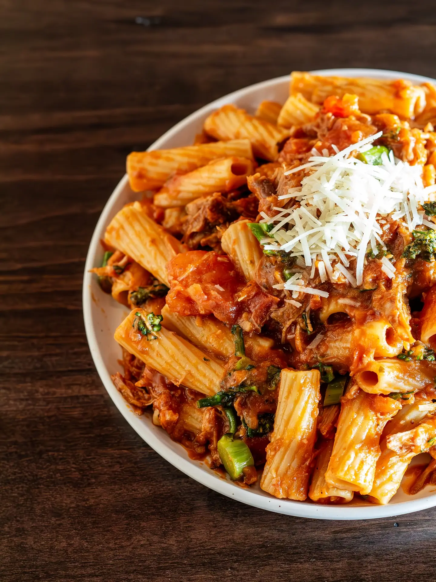 Rigatoni pasta in chunky tomato sauce with shredded meat, greens and grated Parmesan on a white plate over a wooden table