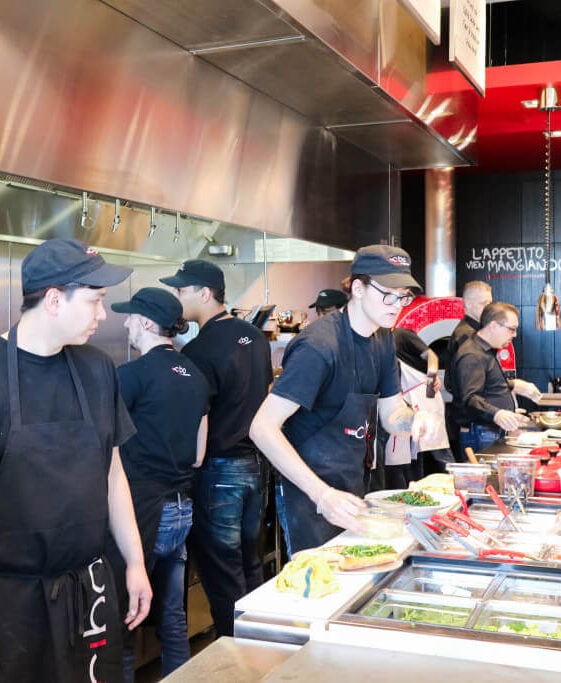 Busy restaurant kitchen line with staff in black shirts and caps preparing plates and assembling orders at a stainless steel counter