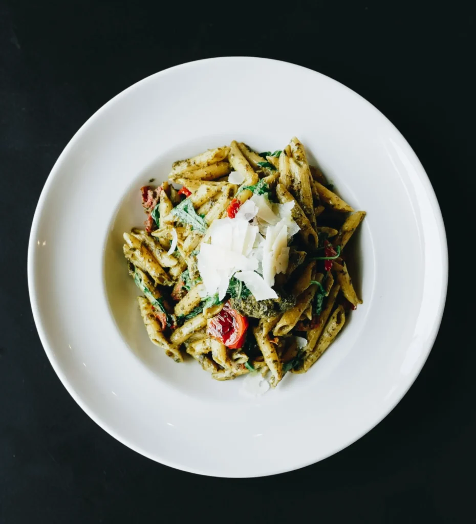 Penne pasta tossed in green pesto with cherry tomatoes, spinach and shaved Parmesan on a white plate over a dark table