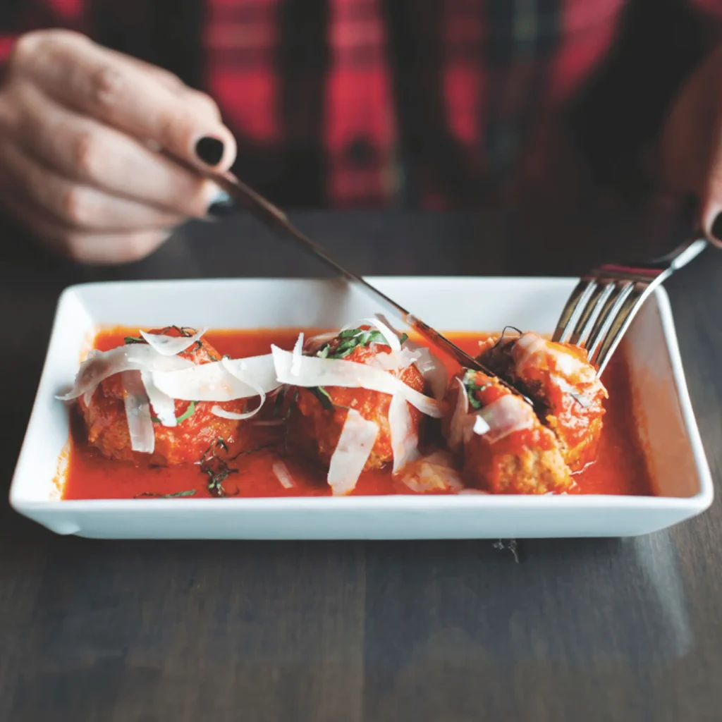 Close-up of meatballs in red tomato sauce topped with shaved cheese, a person cutting a bite with a fork and knife.