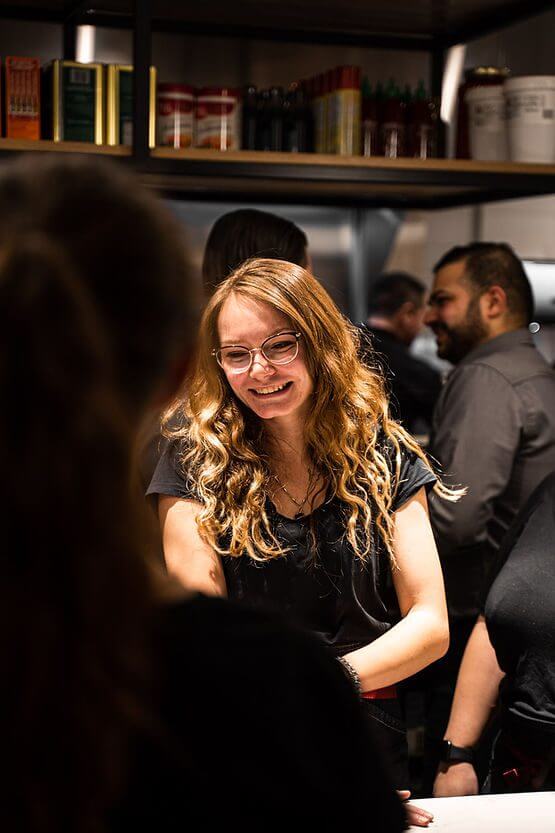 Young woman with wavy blonde hair and glasses smiling while interacting with people in a busy indoor setting.