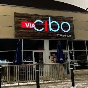 Entrance of a Via Cibo Italian street food restaurant at night with illuminated red and blue sign, outdoor umbrellas and metal railing in front.